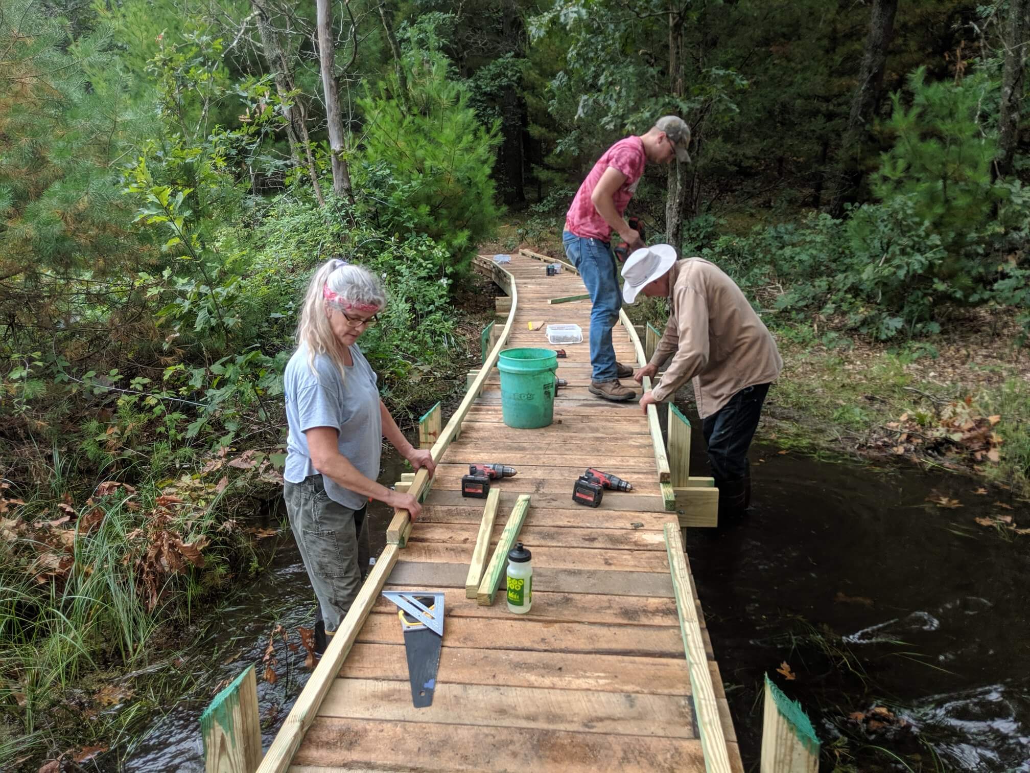 Three volunteers with drills and saws installing a new wooden boardwalk over a creek at a Land Trust preserve