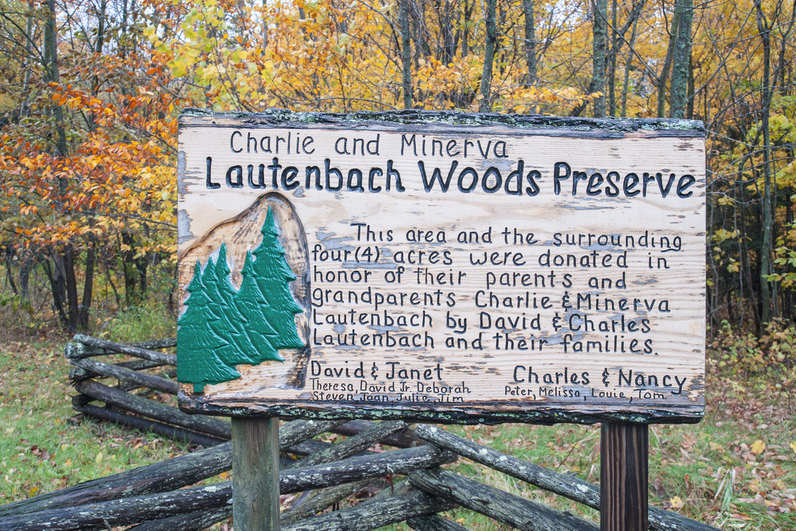 A hand-carved wooden sign reading Charlie and Minerva Lautenbach Woods Preserve stands behind a split-rail fence framed by golden autumn foliage.