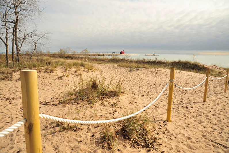 A rope-and-post fence curves across soft sand and dune grass toward the distant red ship canal lighthouse and pier under an overcast sky.