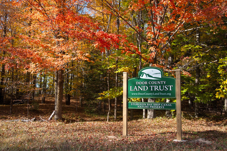 A Door County Land Trust preserve sign stands at a leaf-covered trailhead surrounded by brilliant orange and red maples in full autumn color.