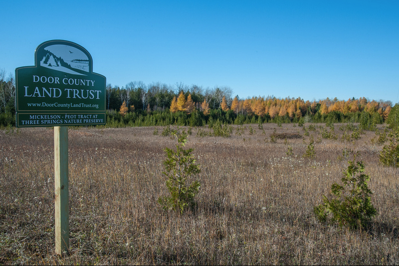 A green Door County Land Trust sign marks the edge of an open meadow backed by yellow tamaracks and mixed hardwoods in fall.