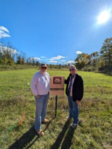 A man and woman stand in an open field proudly next to a sign designating their land as a conservation easement, protected forever for conservation. 