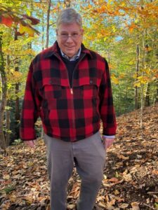 A man in a bright red plaid shirt smiles as he stands under orange-colored fall foliage
