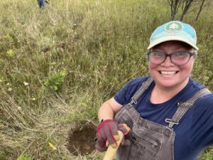 The author Carrie smiles a big smile at the camera as she takes a selfie with the hole she has dug on Washington island, where a shrub will be planted to benefit birds.