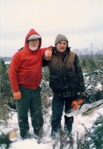 Two men with their arms over each others shoulders stand in a snowy open field, the younger man looks tired and holds a chainsaw.