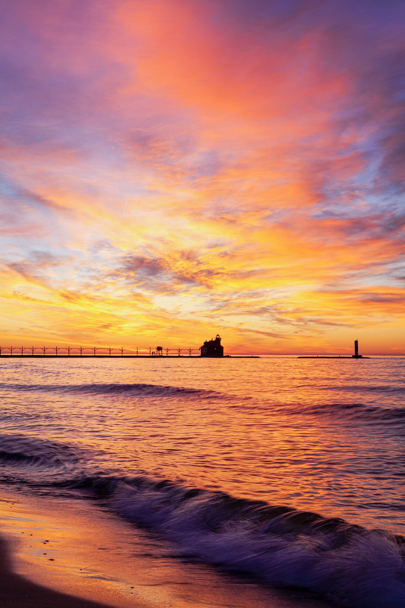 A vivid sunset glowing over the water with a pier and lighthouse in silhouette